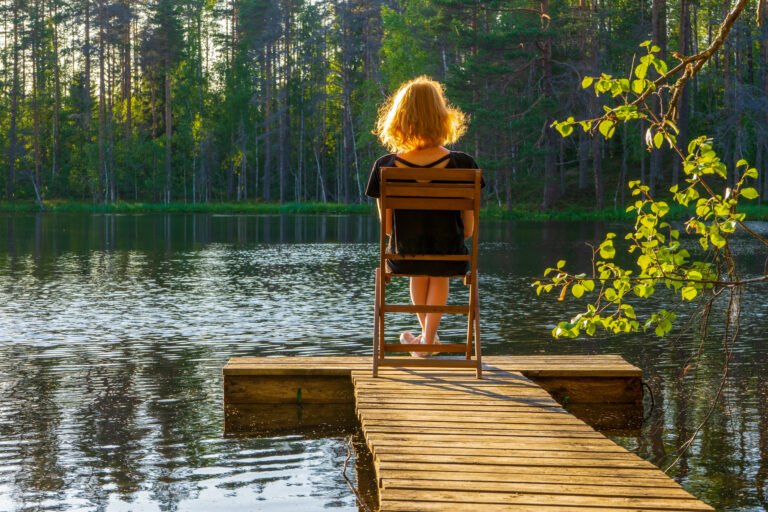 Rear view of young redhead woman sitting on chair on wooden planked footway at lake in summer evening at sunset. Beautiful girl relaxing against northern landscape of pond and forest. Leisure concept