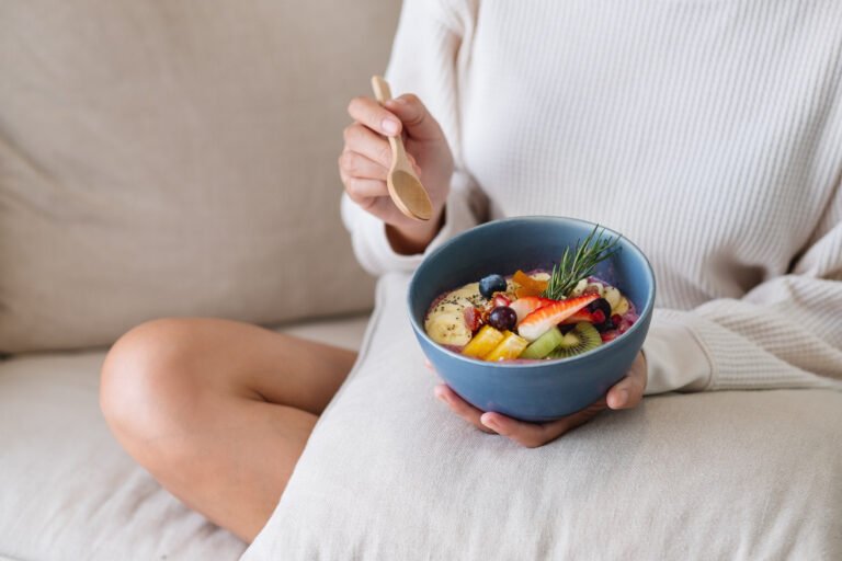A woman eating healthy blueberry smoothie bowl with mixed fruits topping on sofa at home