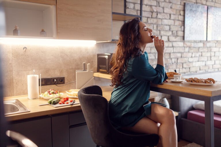 Pretty brunette woman in silk robe biting the cookie while enjoying coffee sitting at the kitchen at home, closed eyes. Morning routine, domestic life