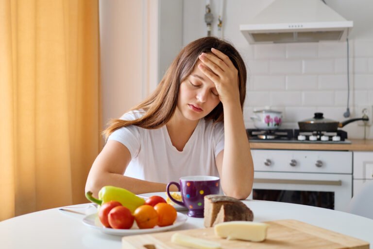 A woman sits at a kitchen table, looking distressed with her hand on her forehead. The table is set with a mug, fruits, and bread, indicating a morning meal