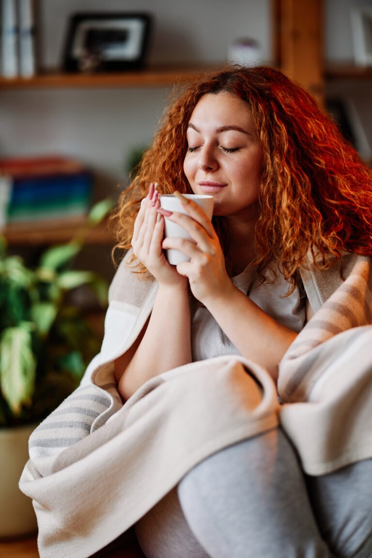 A beautiful ginger girl sitting in the chair in the living room wrapped in a blanket and smelling her morning coffee.