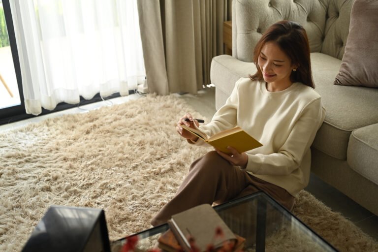 Pretty young woman sitting in cozy living room and making notes to her diary.