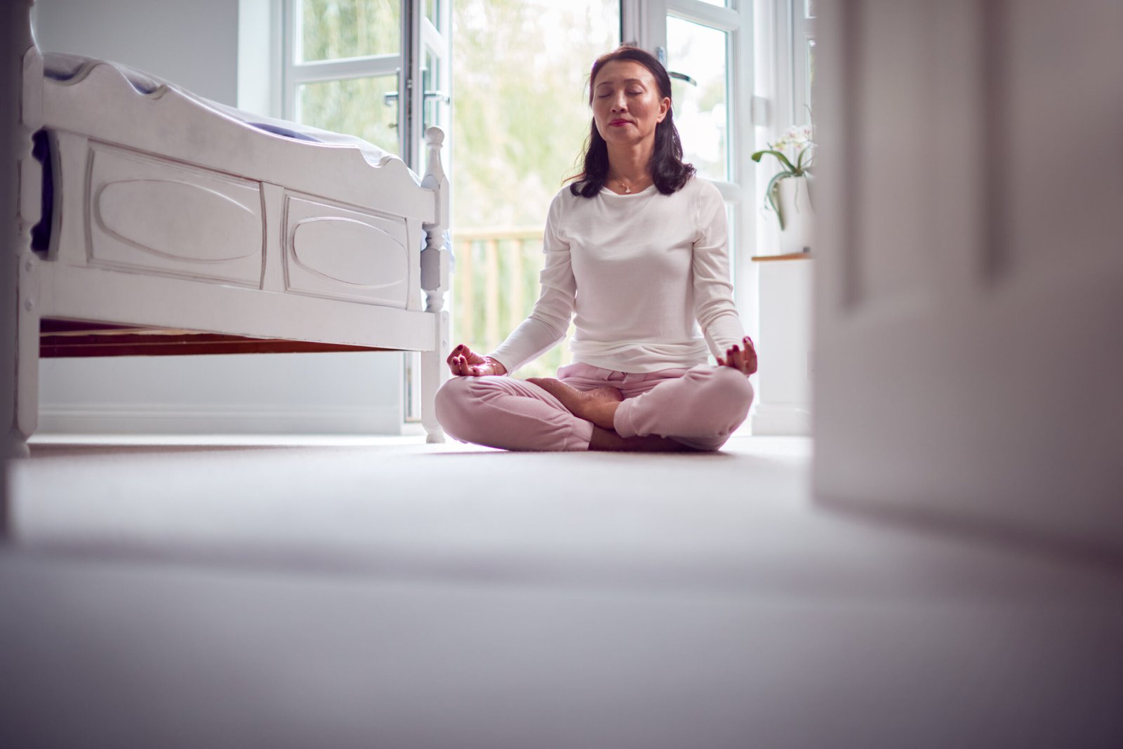 Mature Asian Woman In Pyjamas Sitting On Bedroom Floor Meditating In Yoga Pose