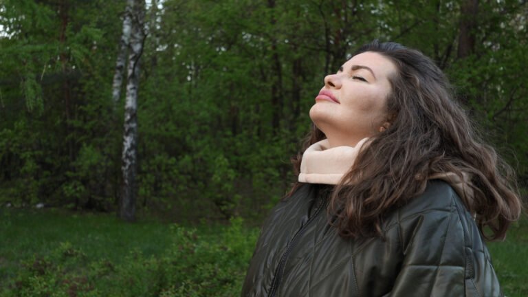 relaxed young woman smiles, breathing fresh forest air deeply amid green trees on serene morning, embracing mindful harmony with nature’s vitality. Concept: Mindful harmony through nature immersion.