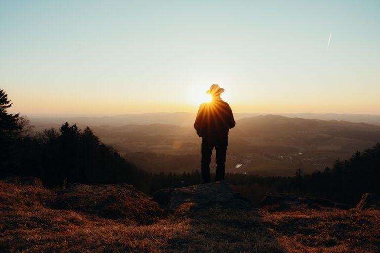 Silhouette of a person in a hat standing on a hilltop at sunset, overlooking a vast landscape