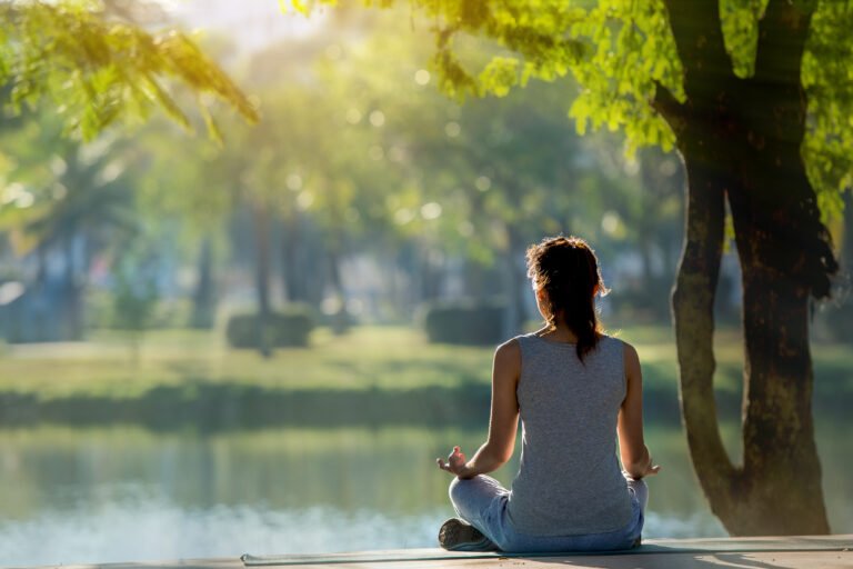 Rear View Of Young Woman Meditating While Sitting By Lake At Park