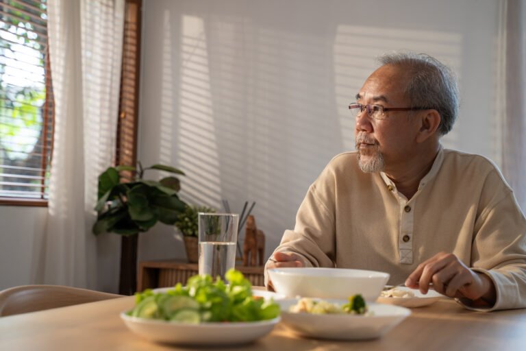 Unhappy Asian Senior older man sit alone, eat foods on table in house. Depressed mature attractive elderly retired grandfather stay at home with painful face feeling upset, lonely and miss his family.