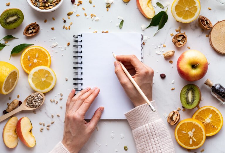 Female hand write in notebook on healthy food background, women diet nutrition recipe menu, fresh summer fruit granola seeds on white table organic super food, health care detox, top view, copy space