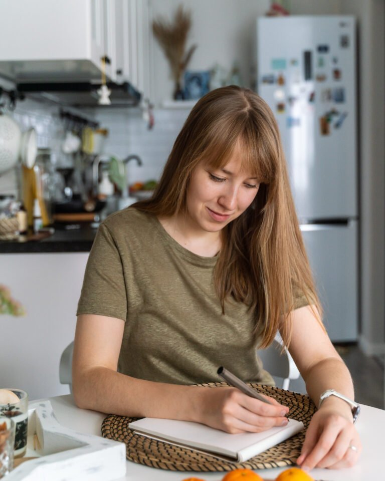 Young female making notes in notebook sitting in kitchen. Planning buying. Making list of products
