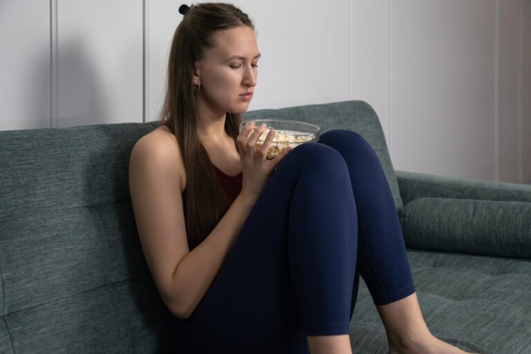 Sad woman sitting on a sofa in her apartment, watching a dull movie while eating popcorn, feeling a deep sense of boredom and loneliness during her weekend leisure time