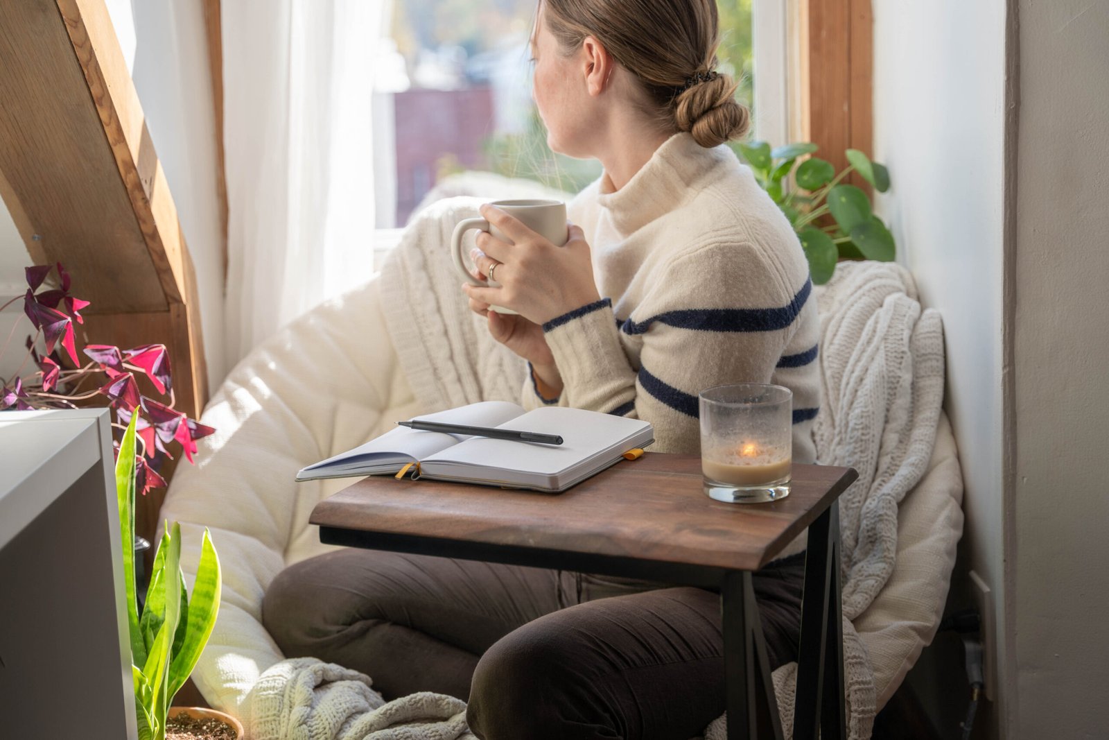Young woman taking a mental health break to write in her journal stares out the window with coffee in hand