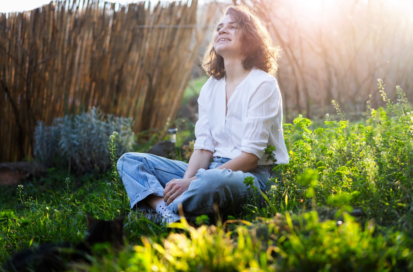 Happy pretty young cheerful woman in white shirt sitting on grass enjoying sunny spring day