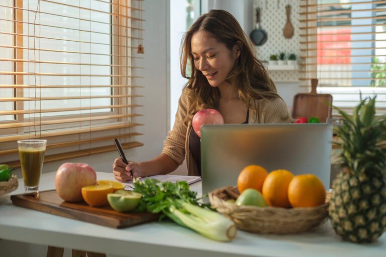 Beautiful young woman sitting in kitchen and writing down healthy recipe or daily ration diet. Healthy lifestyle concept.
