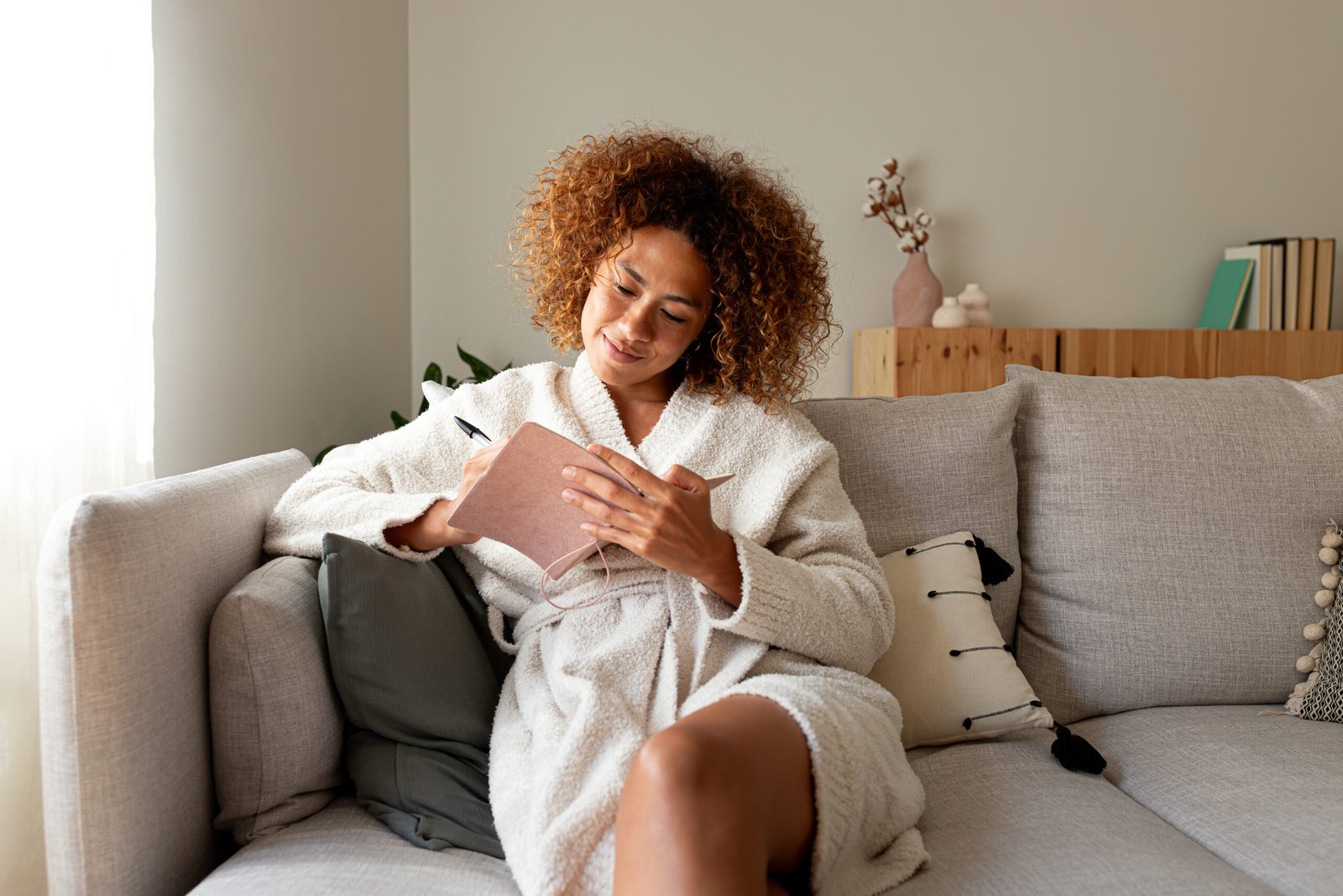 Multiracial woman writing on personal journal sitting on couch at home. African American female writing notes with pen.