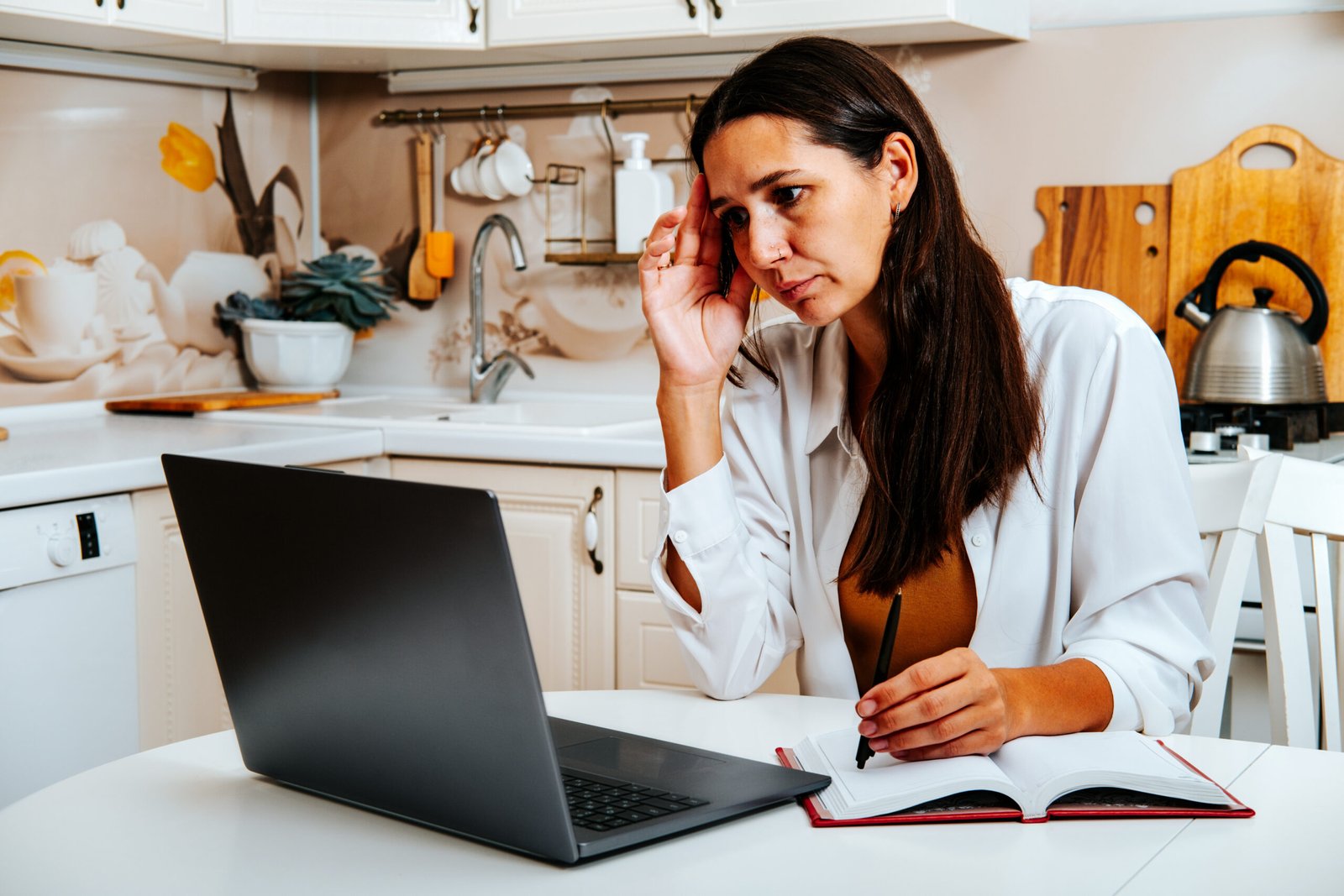 Portrait of female student watching webinar or tutorial studying online on laptop sitting at kitchen table, noting down after teacher or lecturer.