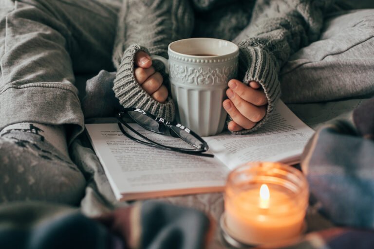 Female hands holding cozy mug with cacao or chocolate. Woman in gray knitted sweater sitting on bed.