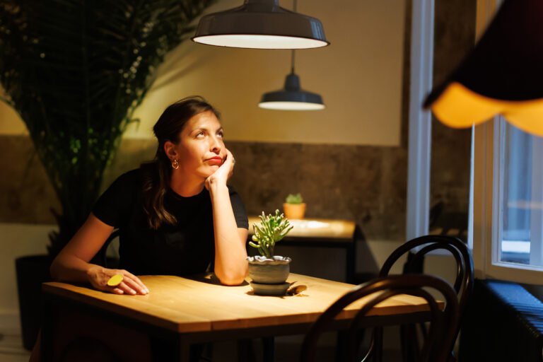 A woman in a black shirt sits at a wooden table, lost in thought, under soft indoor lighting in a tranquil cafÃ© setting
