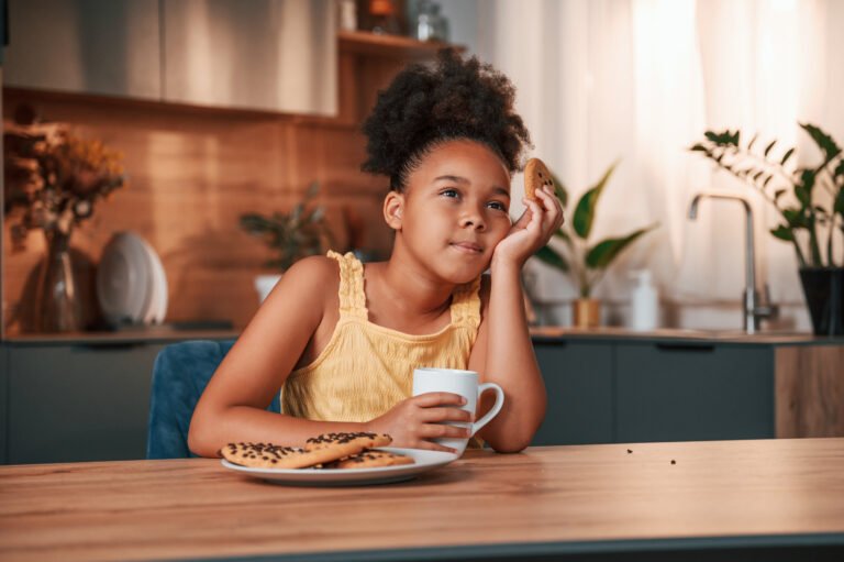 Eating cookies and holding cup. Cute black girl in casual clothes is at home at daytime