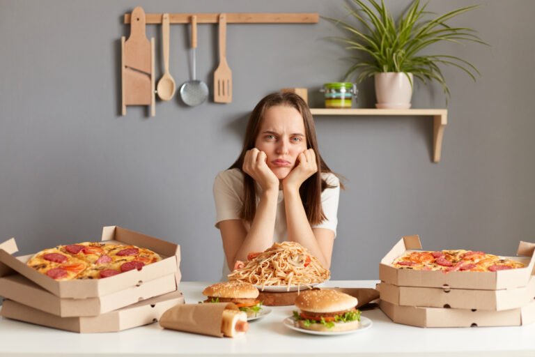 Horizontal shot of upset bored Caucasian woman wearing white casual T-shirt sitting at table in kitchen, keeps hand under chin, looking at junk food, can't eat, keeps diet.