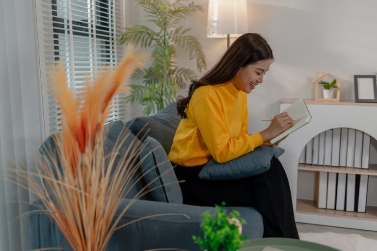 Young Asian woman in yellow sweater sitting on sofa and smiling while writing notes in notebook. Relaxed atmosphere in cozy living room. Concept of learning, planning, journaling, or creative thinking